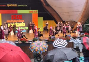 Na pierwszym planie grupa osób z otwartymi parasolkami a w tle na scenie tańczące pary w gruzińskich strojach ludowych