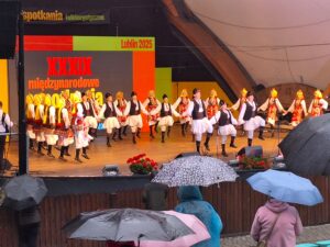 Na pierwszym planie grupa osób z otwartymi parasolkami a w tle na scenie tańczące pary w strojach ludowych