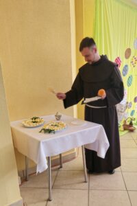 Stojący przy stole mężczyzna w brązowym habicie święcący kropidłem pokrojone w ćwiartki jajka ułożone na tacach na białym obrusie