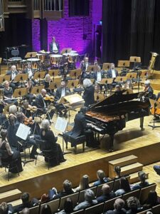 Fragment wnętrza sali w filharmonii z widownią i widzami oraz sceną z członkami orkiestry i instrumentami