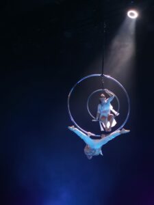 Kobieta i mężczyzna ubrani na biało i wykonujący akrobacje na zawieszonych wysoko dwóch metalowych kołach