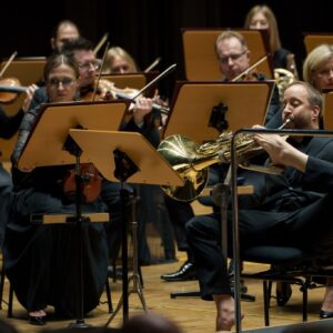 Część siedzących i grających na instrumentach muzyków orkiestry a wśród nich mężczyzna grający lewą stopą na waltorni