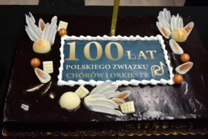 Dekoracyjny tort w kształcie prostokąta z napisem 100 lat Polskiego Związku Chórów i Orkiestr