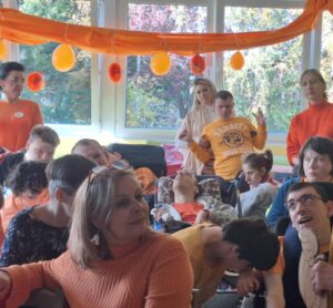 Dorosłe osoby w pomarańczowych ubraniach siedzące na krzesłach ustawionych w rzędach