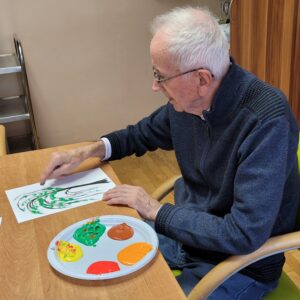 Dorosły mężczyzna siedzący przy stole i malujący palcami jesienne drzewo na białej kartce