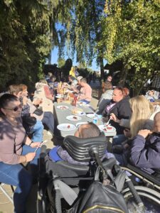 Duża ilość osób siedzących i jedzących przy stole ustawionym wśród drzew i zieleni