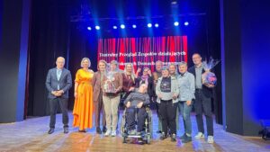 Dwanaście dorosłych osób stojących na scenie oświetlonej na niebiesko i czerwono