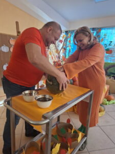 Dwie dorosłe osoby w pomarańczowych ubraniach trzymające dłonie na dyni leżącej na stoliku z pomarańczowym obrusem