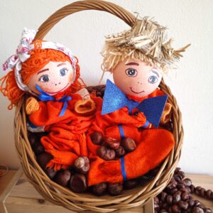 Dwie kukiełki w czerwono-niebieskich kolorach siedzące w wiklinowym koszyku wypełnionym kasztanami