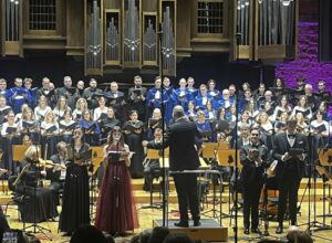 Dyrygent stojący tyłem do widza oraz kilka osób siedzących i trzymających instrumenty a w tle liczna grupa stojących w rzędach osób pod instrumentem organów