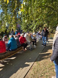 Gruba osób w kurtkach siedząca na dwóch długich ławach ustawionych pod drzewem z zielonymi liśćmi