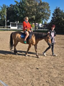 Kobieta siedząca w siodle  na brązowym koniu prowadzonym przez instruktora