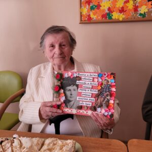 Kobieta w białym żakiecie siedząca przy stole i trzymająca w dłoniach laurkę z umieszczonymi na niej dwoma zdjęciami i życzeniami