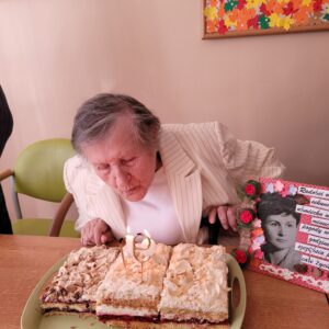 Kobieta w białym żakiecie siedząca przy stole i zdmuchująca świeczkę umieszczoną na kolorowym cieście