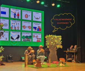 Na scenie z leśną scenografią siedzące dwie dorosłe osoby w kapeluszach oraz w tle na ścianie wyświetlone piktogramy