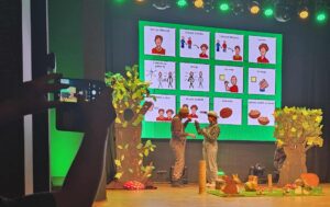 Na scenie z leśną scenografią stojących dwoje dorosłych w kapeluszach oraz w tle na ścianie wyświetlone piktogramy