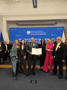 Na tle niebieskiego telebimu grupa kobiet i mężczyzn prezentująca pamiątkowy list gratulacyjny