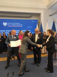 Na tle niebieskiego telebimu i grupy dorosłych osób widzimy dwie kobiety podające sobie dłonie