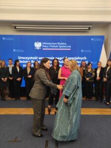 Na tle niebieskiego telebimu i grupy kobiet i mężczyzn widzimy dwie kobiety podające sobie dłonie