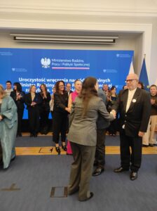 Na tle niebieskiego telebimu i grupy kobiet i mężczyzn widzimy kobietę i mężczyznę podających sobie dłonie