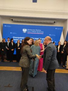 Na tle niebieskiego telebimu i grupy stojących osób widzimy kobietę i mężczyznę podających sobie dłonie