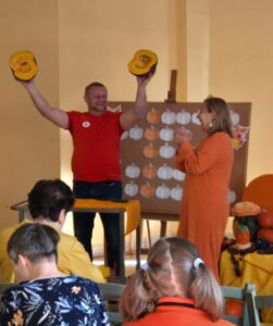 Osoby siedzące na krzesłach a w tle mężczyzna w pomarańczowym podkoszulku trzymający dwie połówki dyni w uniesionych rękach