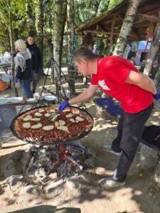 Pochylony mężczyzna w czerwonym podkoszulku stojący nad okrągłym rusztem zawieszonym na łańcuchach nad paleniskiem