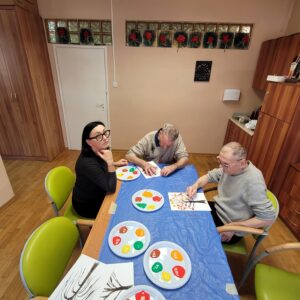 Trzy dorosłe osoby siedzące przy stole nakrytym niebieską ceratą i malujące palcami jesienne drzewa na białych kartkach