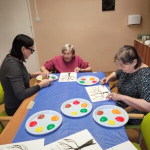 Trzy kobiety siedzące przy stole nakrytym niebieską ceratą i malujące palcami jesienne drzewa na białych kartkach