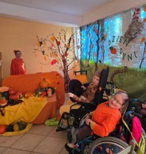 We wnętrzu dwie osoby na wózkach inwalidzkich i jedna stojąca w jesiennej scenografii w pomarańczowych kolorach