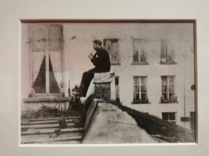 Czarno-biała fotografia przedstawiająca mężczyznę siedzącego na kominie i grającego na flecie