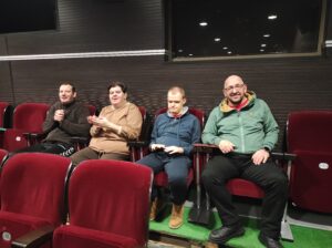 Cztery dorosłe osoby siedzące w ciemnoczerwonych fotelach widowiskowych