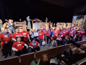 Duża grupa dorosłych osób przeważnie w czerwonych podkoszulkach stojących na scenie na tle dekoracji imitującej wiejską zabudowę