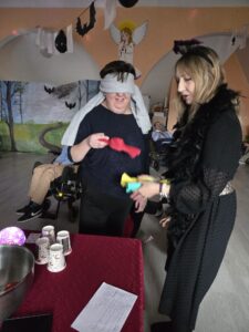 Dwie kobiety w czarnych strojach z których jedna ma zasłonięte oczy białym materiałem i trzyma w dłoniach przedmiot przypominający duży czerwony cukierek