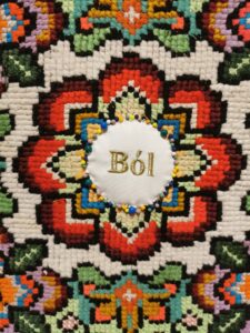 Fragment kolorowej wyszywanki w formie geometrycznego kwiatu ze słowem ból wyszytym na środku złotymi literami