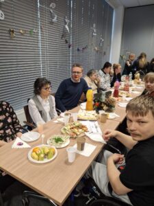 Grupa dorosłych osób siedzących przy brązowym stole zastawionym słodkim poczęstunkiem na białych talerzach