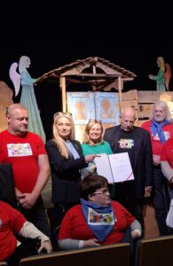 Grupa dorosłych osób trzymających dyplom i stojących na scenie na tle dekoracji imitującej wiejską zabudowę