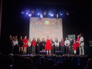 Grupa młodych osób stojących w półkolu na oświetlonej scenie na tle wyświetlonej kolorowej grafiki przedstawiającej Jezusa w stajence