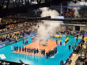 Hala sportowa z widzami a na środku boisko do piłki siatkowej dookoła którego stoją zwodnicy drużyn biorący udział w meczu