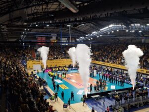 Hala sportowa z wypełnionymi trybunami oraz z zawodnikami drużyn siarkowych i członkami górniczej orkiestry stojącymi wzdłuż linii boiska