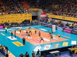 Hala sportowa z wypełnionymi trybunami oraz zawodnikami rozgrywającymi na boisku mecz piłki siatkowej