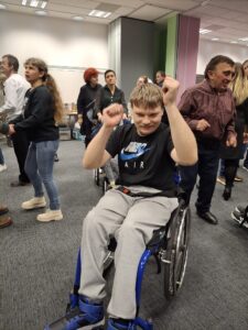 Kilka par dorosłych osób stojących na szarej wykładzinie a na pierwszym planie osoba na wózku inwalidzkim z podniesionymi rękami zgiętymi w łokciach