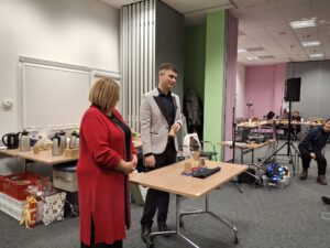 Kobieta i mężczyzna stoją za małym stolikiem na którym znajdują się trzy papierowe kubeczki i pionowy szpikulec między nimi