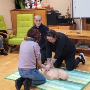 Kobieta instruowana przez mężczyznę i kobietę wykonuje uciski okolicy międzymostkowej na fantomie