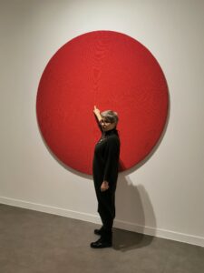 Kobieta w czarnej sukience dotykająca środka dużego czerwonego koła wiszącego na białej ścianie