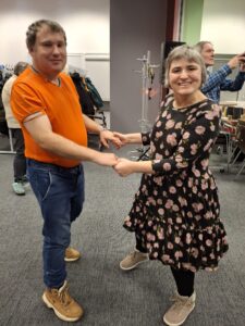 Kobieta w czarnej sukience w różowe kwiaty i mężczyzna w pomarańczowym podkoszulku tańczą trzymając się za ręce