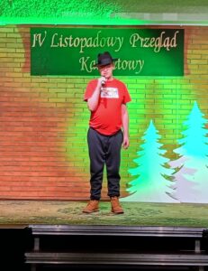 Mężczyzna w czerwonym podkoszulku i kapeluszu trzymający mikrofon i stojący na scenie oświetlonej zielonym światłem