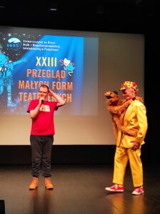 Mężczyzna w czerwonym podkoszulku trzymający mikrofon i mężczyzna ubrany na żółto stoją na tle projekcji na scenie