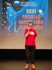 Mężczyzna w czerwonym podkoszulku trzymający mikrofon i stojący na tle projekcji na scenie