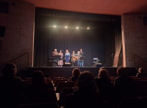 Sala widowiskowa z widzami w fotelach oraz sześcioma osobami stojącymi na scenie z keyboardem gitarą i mikrofonami